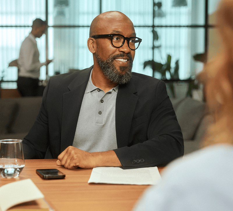 Man in a suit sitting at a table with a woman, engaged in conversation, in an office setting.